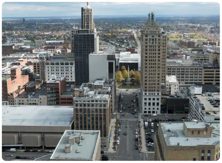 Aerial view of a city with several tall office buildings, streets lined with cars, and a mix of modern and historic architecture under a cloudy sky.